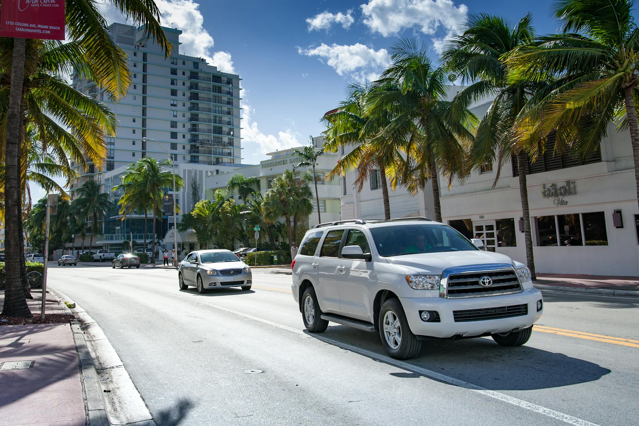 A vibrant city street lined with palm trees and modern buildings under a bright sky.
